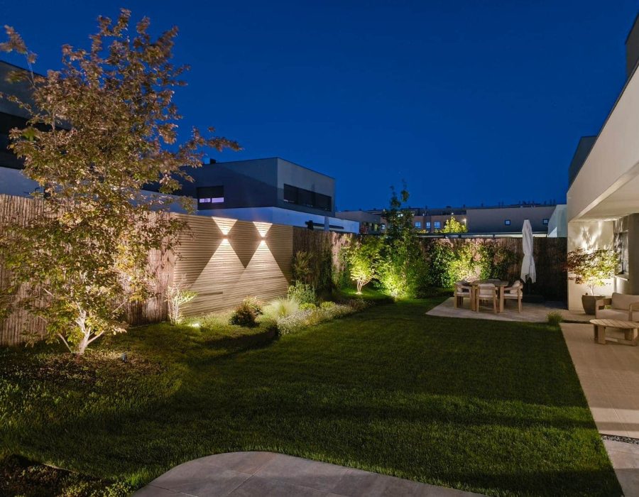Vista nocturna de jardín con césped, iluminación de arbolado y comedor exterior en Corredor del Henares — Abierto al Jardín