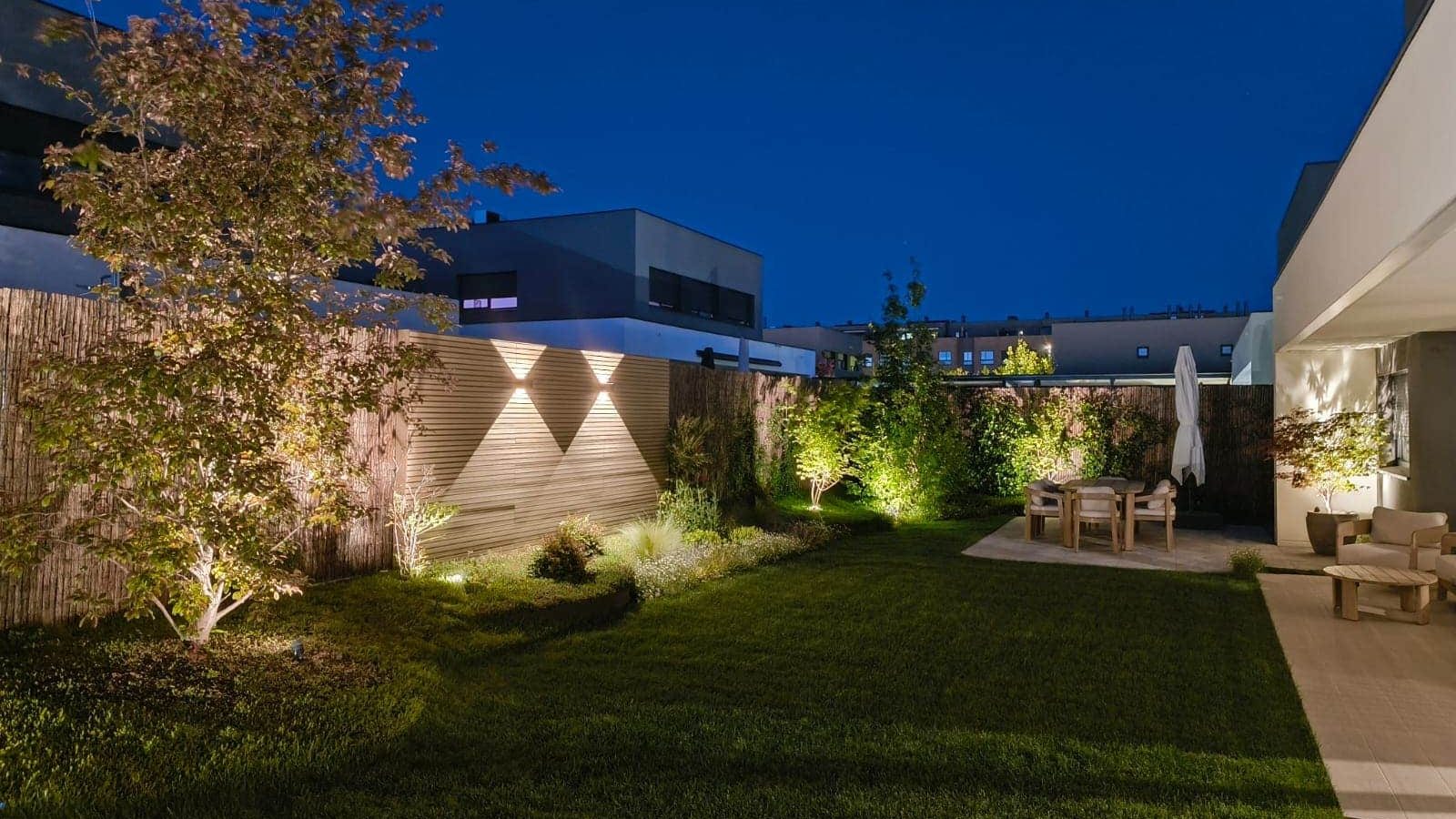 Vista nocturna de jardín con césped, iluminación de arbolado y comedor exterior en Corredor del Henares — Abierto al Jardín